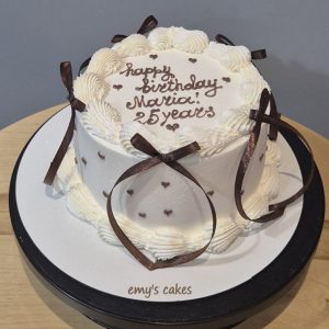 Tort alb acoperit în cremă, fundițe maro, mesaj Happy Birthday și inimioare