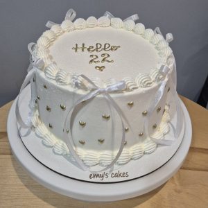 Tort alb cu cremă de brânză, inimioare aurii și fundițe albe, mesaj „Hello 22”