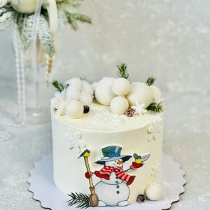 Tort de iarnă acoperit în cremă de brânză, decorat cu bulgări albi, crenguțe de brad și om de zăpadă desenat