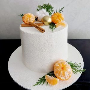 Tort înalt alb cu efect catifelat, decorat cu mandarine, baton de scorțișoară, rămurele verzi, glob auriu și bumbac