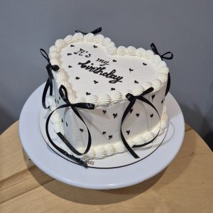 Tort în formă de inimă, acoperit în cremă de brânză albă, cu fundițe negre și inscripție „It’s my birthday”, realizat artizanal de Emys Cakes.