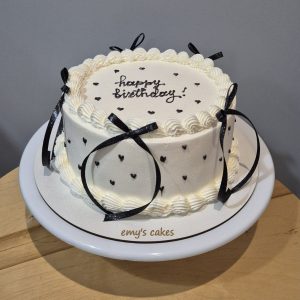 Tort rotund acoperit în cremă de brânză albă, decorat cu fundițe negre și inimioare mici, cu inscripția „happy birthday”, realizat artizanal de Emys Cakes.