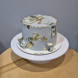Tort rotund acoperit în cremă de brânză gri deschis, decorat cu teancuri de dolari comestibili, bancnote rulate și detalii aurii, cu topper „La Mulți Ani”, realizat de Emys Cakes.