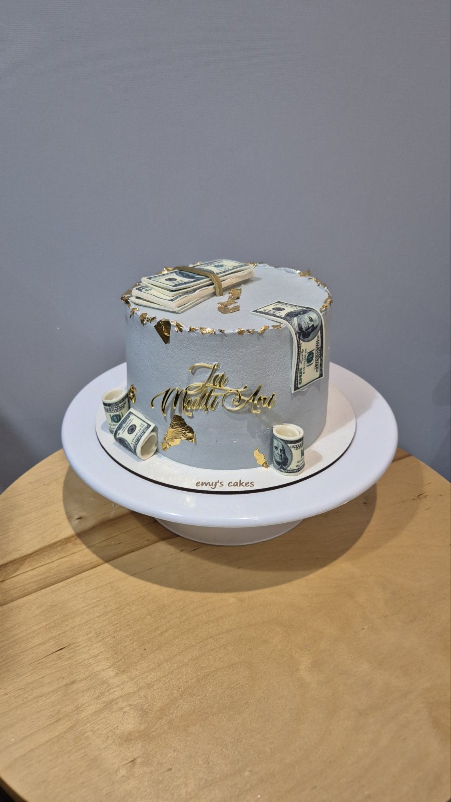 Tort rotund acoperit în cremă de brânză gri deschis, decorat cu teancuri de dolari comestibili, bancnote rulate și detalii aurii, cu topper „La Mulți Ani”, realizat de Emys Cakes.