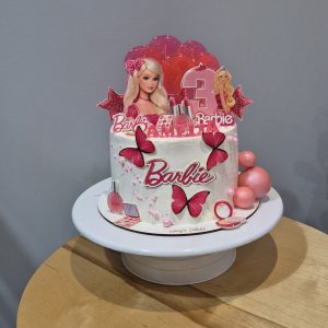 Tort Barbie pentru fetițe, acoperit în cremă albă cu accente roz, decorat cu personaje Barbie, fluturi roz, bile lucioase și cifra 3, realizat de Emys Cakes.
