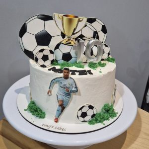 Tort tematic fotbal acoperit în cremă de brânză albă, decorat cu mingi de fotbal, trofeu auriu, cifra 10 argintie și jucător în acțiune, realizat de Emys Cakes.