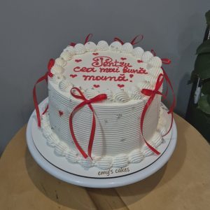 Tort alb acoperit în cremă, decorat cu fundițe roșii, perle argintii și mesaj „Pentru cea mai bună mamă!”, realizat de Emys Cakes.