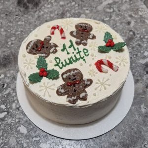 Bento cake alb de iarnă, decorat cu omuleți de turtă dulce, bastonașe „candy cane”, frunze verzi cu bobițe roșii și mesaj „11 lunițe”, realizat de Emys Cakes.