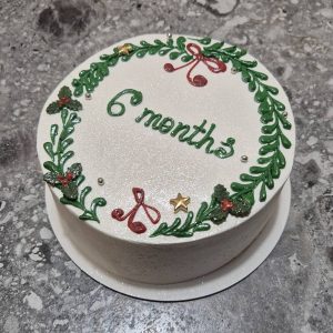 Bento cake alb sidefat, decorat cu coroniță verde de Crăciun, bobițe roșii, fundițe și steluțe aurii, cu mesaj „6 months”, realizat de Emys Cakes.