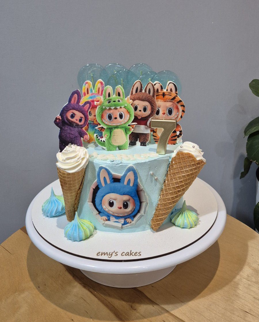 Tort pentru copii acoperit în cremă de brânză albastru pastel, decorat cu personaje haioase în costume de animăluțe, conuri de înghețată, bezele colorate și lumânare cu cifra 7, realizat de Emys Cakes.