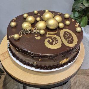Tort aniversar mare, glazurat în ganaj de ciocolată, decorat cu bile aurii, inscripția „50” și topper „La Mulți Ani”, realizat de Emys Cakes.