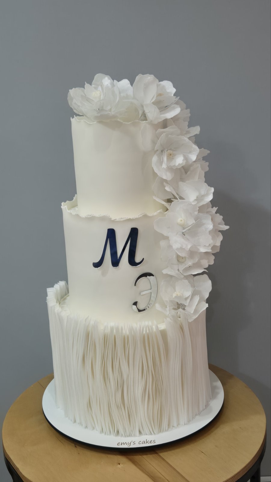 Tort nuntă elegant acoperit în pastă de zahăr albă, cu flori albe și inițiale M&E, decor premium Emys Cakes