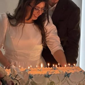 Tort de nuntă acoperit în cremă, cu decor floral albastru și lămpi calde, cu miri în fundal, Emys Cakes