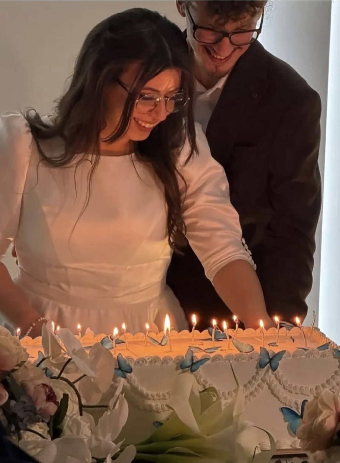 Tort de nuntă acoperit în cremă, cu decor floral albastru și lămpi calde, cu miri în fundal, Emys Cakes